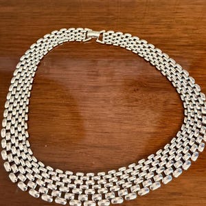Puede incluir: Collar plateado con diseño de múltiples hilos. El collar presenta una serie de eslabones rectangulares conectados para formar un collar. El cierre es visible en la parte superior. El collar se muestra sobre una superficie de madera.