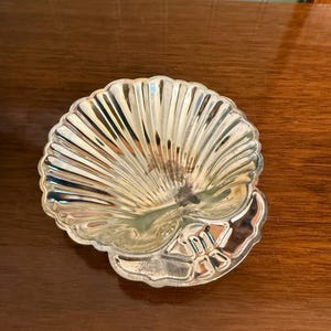 May include: A silver-toned metal dish shaped like a scallop shell. The dish features a ribbed, fan-like interior surface with a smooth, curved base section. The metallic finish is reflective with visible signs of tarnish and wear in the center.