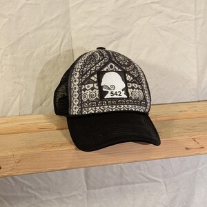 May include: Black trucker hat with a patterned design on the front panel. The design is black and white, featuring a silhouette and the number 542. The hat has a black brim and mesh back.