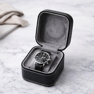 Puede incluir: Estuche de reloj de cuero negro con costuras blancas, abierto para revelar un reloj con esfera negra y correa de cuero negro. El estuche tiene un interior gris y está sobre una superficie de mármol.