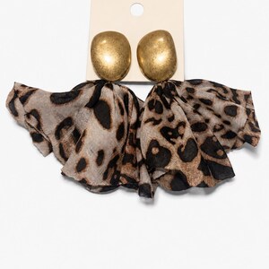 May include: A pair of statement earrings featuring smooth, brushed gold-tone metal studs attached to large, ruffled fabric drapes. The fabric displays a brown, tan, and black leopard print pattern, creating a voluminous, layered appearance.
