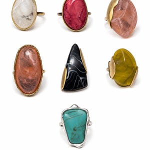 May include: A collection of seven rings featuring various shapes and colors, including clear, red, pink, black, olive green, and turquoise stones set in gold-toned and silver-toned metal bands, arranged in a grid against a plain white background.