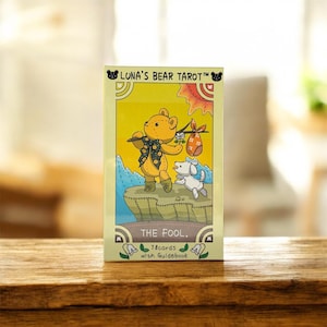 以下が含まれることがあります： 「Luna's Bear Tarot」というタイトルのタロットカードデッキ。黄色いクマのキャラクターが杖を持ち、白い犬が描かれています。カードには「THE FOOL」と書かれており、「78枚のカードとガイドブック」が含まれています。背景は山と太陽のデザインの黄色です。
