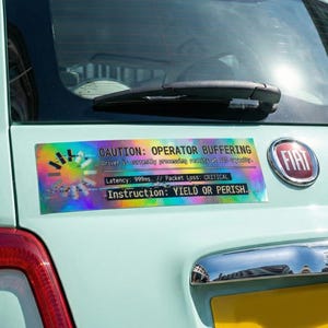 Puede incluir: Una pegatina holográfica en un coche azul claro con el texto "CAUTION: OPERATOR BUFFERING" y otros datos técnicos. La pegatina también incluye la instrucción "YIELD OR PERISH". Se ve un emblema de Fiat.