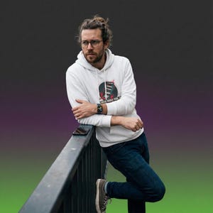 Puede incluir: Una sudadera con capucha blanca con un diseño gráfico que presenta un emblema circular y texto. La persona lleva gafas y un reloj inteligente, apoyada en una barandilla negra. La persona lleva vaqueros azules y zapatos grises.