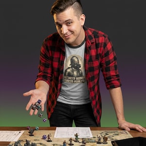May include: A person wearing a gray t-shirt with the text "OUTDATED ARMOUR MODERN PROBLEMS" and a red and black plaid shirt. The person is holding dice above a tabletop game with miniature figures and a map.