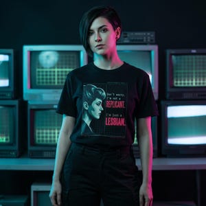 May include: Black t-shirt featuring a side profile of a woman with the text "Don't worry, I'm not a REPLICANT. I'm just a LESBIAN." The graphic is in a grid-like design with a retro aesthetic. The person is wearing black pants.