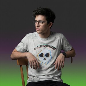 Puede incluir: Camiseta gris claro con un gráfico de calavera pixelada y la palabra "CONSCIOUSNESS". La camiseta tiene un efecto marmoleado y el texto "ERROR 404" encima de la calavera. La persona lleva gafas y un reloj negro.