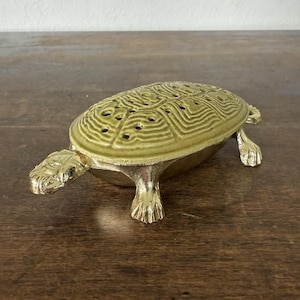 May include: A turtle-shaped decorative box with a shiny gold-toned metal base and legs. The removable lid is made of a textured, pale olive-green ceramic material featuring a detailed shell pattern with small ventilation holes across the surface.