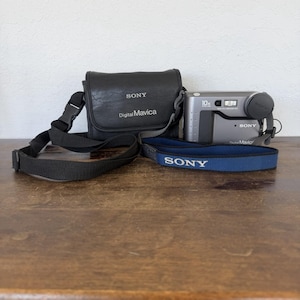 May include: A vintage Sony Digital Mavica camera kit. The kit includes a silver camera with a 10x zoom lens, a black leather carrying case, and a blue and black camera strap. The camera and case have the Sony logo.