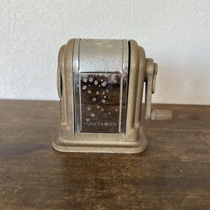 May include: Vintage brass-toned pencil sharpener with a silver metal front panel. The sharpener has a crank handle on the side and the text "PUSH TO OPEN" is visible on the front. The sharpener is sitting on a wooden surface.