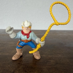 May include: A plastic toy cowboy figure with a white hat, red neckerchief, and blue pants. The cowboy is holding a yellow lasso. The figure is standing on a brown surface.