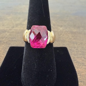May include: A gold-toned ring featuring a large, faceted, rectangular-cut pink gemstone. The ring has a gold band with a gold and silver accent on each side of the gemstone. The ring is displayed on a black velvet ring holder.
