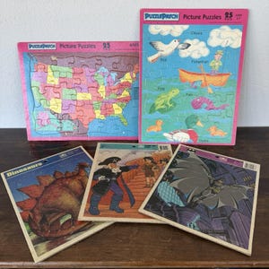 May include: A collection of vintage children's jigsaw puzzles. The puzzles feature colorful illustrations of a map of the United States, dinosaurs, a pirate scene, Batman, and a scene with a fisherman, birds, and ducks. Each puzzle is labeled "PuzzlePatch Picture Puzzles".