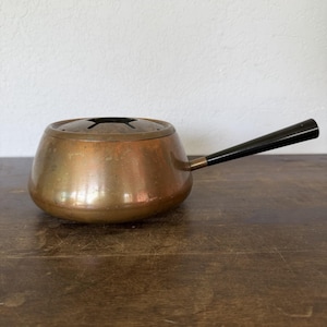 May include: A round copper fondue pot with a smooth, metallic finish and a long, tapered black handle. The top of the pot features a circular opening with a cross-shaped metal insert, designed for holding fondue forks securely in place during use.