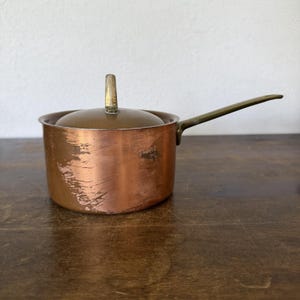 May include: A copper saucepan with a matching lid and a long brass handle. The exterior of the pot shows visible surface wear and scratches, while the lid features a central brass knob. The metal surfaces exhibit a warm, metallic finish.