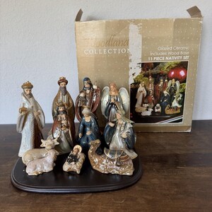 May include: An 11-piece glazed ceramic nativity set from the Woodland Collection, featuring figurines of figures, an angel, and animals on a dark wood base. The original cardboard box is included, displaying the set's contents and product details.