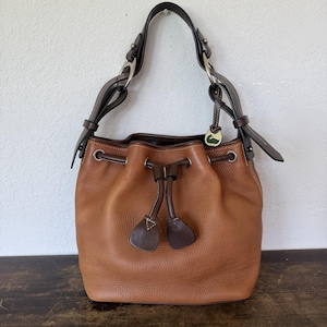 May include: A tan pebbled leather bucket bag featuring a dark brown leather drawstring closure with two matching leather pulls, a single dark brown shoulder strap with silver-tone hardware, and a small duck-shaped metal charm attached to the strap.
