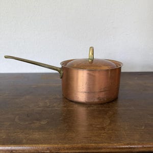 May include: A round copper saucepan with a polished brass handle and a matching copper lid featuring a brass knob. The exterior of the pan shows signs of wear and light surface scratches, with a warm metallic finish throughout the body.