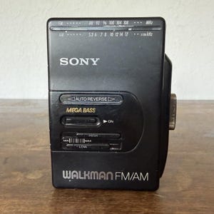 May include: A black Sony Walkman FM/AM portable cassette player featuring an auto reverse function, a Mega Bass switch, and a volume slider with min and max settings. The front panel displays radio frequency scales for FM and AM bands in white text.