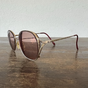 May include: Vintage gold-tone sunglasses with a rounded square frame and tinted lenses. The temples are thin with a red accent at the ear. The sunglasses are resting on a wooden surface.