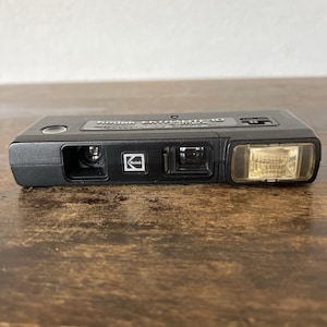 May include: A black Kodak Extralite 10 camera. The camera has a rectangular shape with a flash on the right side. The Kodak logo is visible on the front. The camera is sitting on a wooden surface.