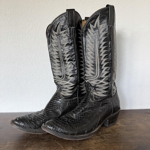 May include: A pair of black western-style boots featuring a textured snakeskin-patterned leather foot and a tall shaft with intricate grey decorative stitching. The boots have a pointed toe, a stacked heel, and pull tabs at the top of the shaft.