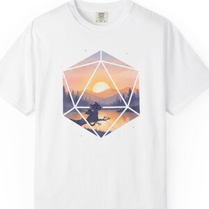 May include: White t-shirt featuring a geometric design with a sunset scene. The design includes a sun, mountains, trees, and a figure on a branch. The shirt has a crew neck and short sleeves.