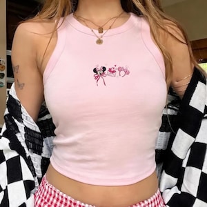 May include: A pale pink vest top with an embroidered Minnie Mouse design, a bow, and hearts. The top has a rounded neckline and a racerback style. The person is wearing a black and white chequered jacket.