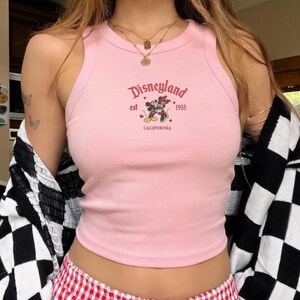 May include: A light pink ribbed tank top with the Disneyland logo, featuring Mickey and Minnie Mouse, the text "est 1955 CALIFORNIA". The top is sleeveless and has a round neckline.