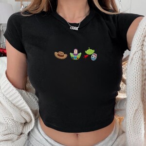 May include: Black crop top with embroidered images of a cowboy hat, Buzz Lightyear, an alien, and a toy character. The shirt has short sleeves and a crew neck.