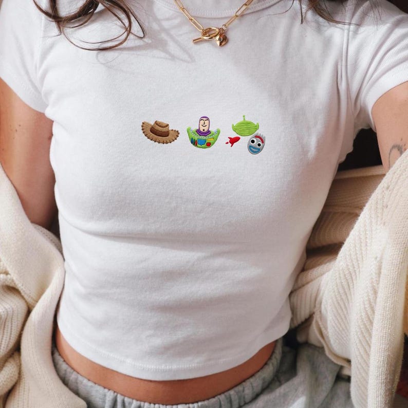 May include: White crop top with embroidered cartoon character designs. The designs include a brown cowboy hat, a green and purple character, a green alien, a red rocket, and a blue and white character.