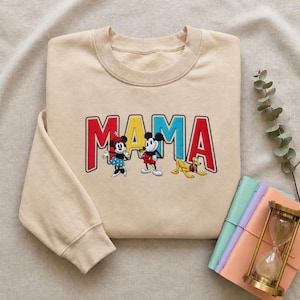 Puede incluir: Sudadera beige con la palabra "MAMA" en letras rojas, amarillas y azules. Minnie Mouse, Mickey Mouse y Pluto est&aacute;n bordados debajo de la palabra. La sudadera est&aacute; doblada sobre una superficie neutra.