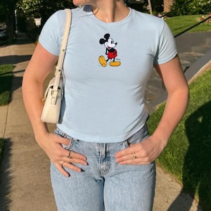 Puede incluir: Camiseta azul claro con un dise&ntilde;o de Mickey Mouse. La parte superior tiene mangas cortas y cuello redondo. El personaje de dibujos animados se muestra con sus cl&aacute;sicos pantalones cortos rojos, zapatos amarillos y guantes negros. La camiseta se combina con vaqueros de mezclilla de lavado claro.