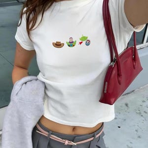 May include: White t-shirt with a small embroidered design featuring a cowboy hat, Buzz Lightyear, a green alien, and a blue alien. The shirt is paired with gray pants, a pink belt, and a red purse.