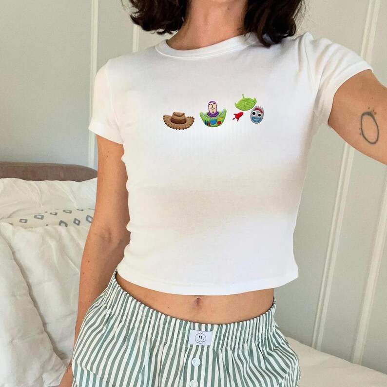 May include: White crew neck t-shirt with embroidered cartoon characters from Toy Story. The shirt features a brown cowboy hat, Buzz Lightyear, a green alien, and a blue and red character. The person is wearing green and white striped pajama pants.