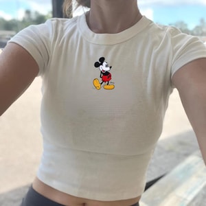 Puede incluir: Top corto de color crema con mangas cortas y una peque&ntilde;a imagen bordada de Mickey Mouse. El personaje de dibujos animados lleva su atuendo cl&aacute;sico: pantalones cortos rojos, zapatos amarillos y guantes blancos. La camiseta tiene cuello redondo.