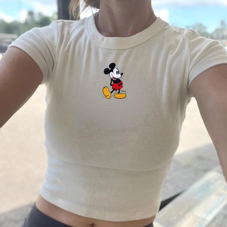 Puede incluir: Top corto de color crema con mangas cortas y una peque&ntilde;a imagen bordada de Mickey Mouse. El personaje de dibujos animados lleva su atuendo cl&aacute;sico: pantalones cortos rojos, zapatos amarillos y guantes blancos. La camiseta tiene cuello redondo.