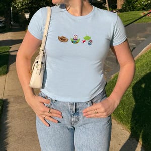 May include: Light blue t-shirt with a crew neck and short sleeves. The shirt features embroidered cartoon character designs. The person is wearing light wash blue jeans and a white purse with a gold chain. The person is also wearing rings.