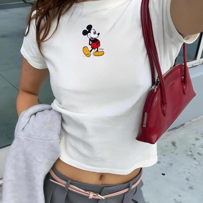 Puede incluir: Camiseta blanca con un dise&ntilde;o de Mickey Mouse. El conjunto incluye pantalones grises, un cintur&oacute;n rosa y un bolso de mano rojo. Un conjunto casual y moderno.
