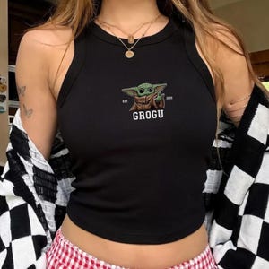 Puede incluir: Camiseta sin mangas negra acanalada con un gr&aacute;fico de Grogu. El gr&aacute;fico incluye una ilustraci&oacute;n verde de Grogu con el texto "EST. 2019" y el nombre "GROGU" en blanco. La camiseta sin mangas es sin mangas y tiene un cuello redondo.