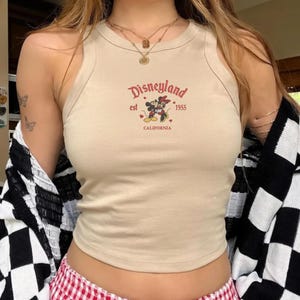 May include: Tan tank top with the Disneyland logo in red, featuring Mickey and Minnie Mouse. The text includes "est 1955 CALIFORNIA". The top is sleeveless and has a round neckline.