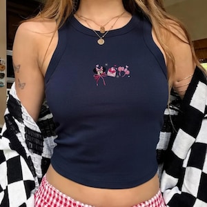 May include: A navy blue vest top with a round neckline. The top has an embroidered design of Minnie Mouse, a pink bow, and a letter. The model is wearing a black and white checkered cardigan and gold necklaces.