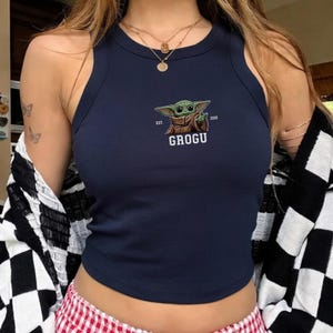 Puede incluir: Top corto azul marino con un gr&aacute;fico de Grogu. La parte superior sin mangas presenta un cuello redondo y una peque&ntilde;a imagen bordada de Grogu con el texto "GROGU" debajo. La parte superior se usa con una chaqueta a cuadros en blanco y negro.