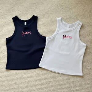 May include: Two sleeveless tank tops. One is navy blue, the other is white. Both have a scoop neck and a small embroidered design in pink and red near the neckline. The tank tops are made of a ribbed material.