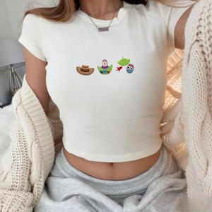 May include: Off-white, short-sleeved crop top featuring embroidered cartoon characters from the movie Toy Story. The design includes a cowboy hat, Buzz Lightyear, a green alien, a rocket, and a blue alien.