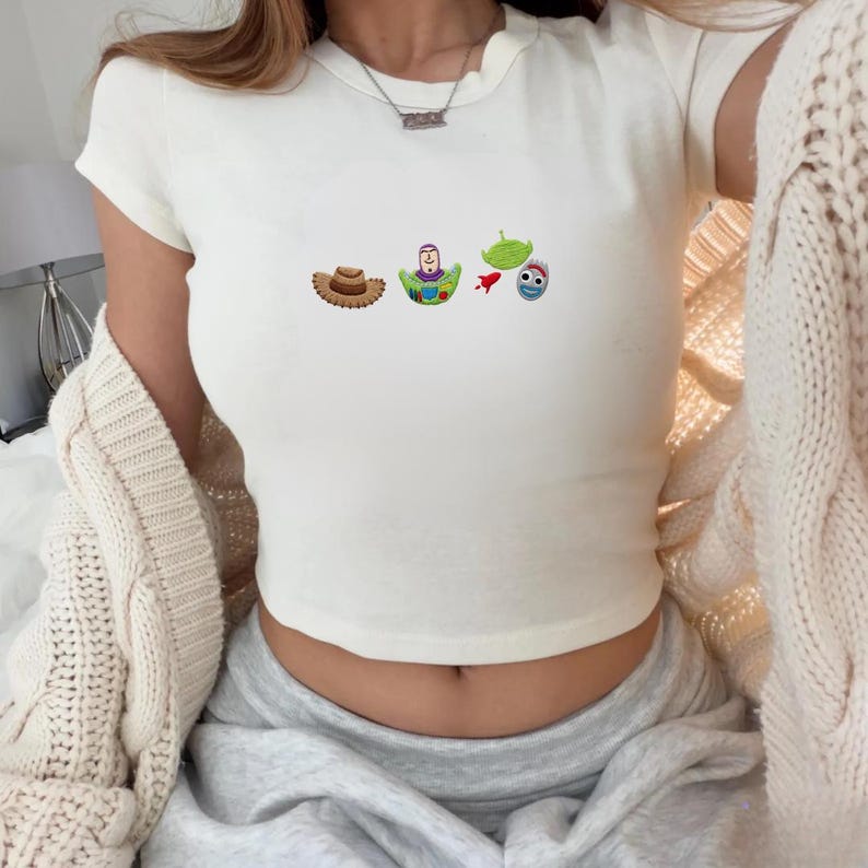 May include: Off-white, short-sleeved crop top featuring embroidered cartoon characters from the movie Toy Story. The design includes a cowboy hat, Buzz Lightyear, a green alien, a rocket, and a blue alien.