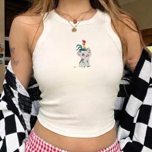 May include: A white crop top with a round neckline. The top features a cartoon illustration of a pig with a rooster on its head. The pig is gray and white, and the rooster is red and yellow. The top is sleeveless.