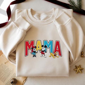 Puede incluir: Sudadera color crema con la palabra "MAMA" en letras rojas, amarillas y azules. La sudadera presenta los personajes de dibujos animados Minnie Mouse, Mickey Mouse y Pluto. Las mangas son largas y con pu&ntilde;os.