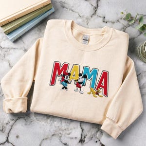 Puede incluir: Sudadera color crema con la palabra "MAMA" bordada en letras rojas, azules y amarillas. La sudadera presenta im&aacute;genes bordadas de Minnie Mouse, Mickey Mouse y Pluto. La sudadera tiene mangas largas y cuello redondo.